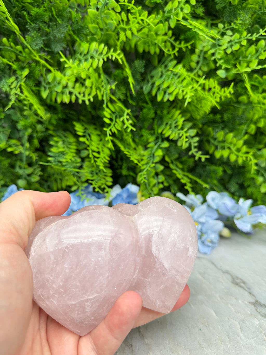 A hand holding a pink crystal against a backdrop of green foliage and blue flowers.