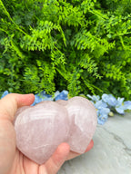 A hand holding a pink crystal against a backdrop of green foliage and blue flowers.