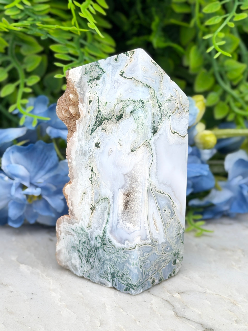 a large, jagged, and textured stone with a mix of white, green, and brown hues, set against a backdrop of lush greenery and blue flowers.